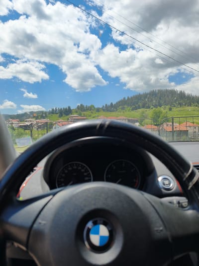 The image is a first-person view from inside a BMW vehicle, likely a passenger car, focused on the steering wheel with the BMW logo. The view through the windshield reveals a rural landscape with a village of Borino, Bulgaria. Buildings with orange tile roofs are visible, along with green fields and a tree-covered hill in the background. The sky is blue with scattered white clouds. Power lines run across the top of the frame. The dashboard is blurred, obscuring details of the speedometer and other controls. No people are visible.