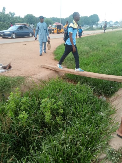 The image depicts a roadside scene in Kagini, Nigeria, under an overcast sky suggesting either early morning or a cloudy day. In the foreground, a young man wearing a blue and yellow shirt, black pants, and a backpack, is walking across a narrow wooden plank that bridges a ditch filled with lush green grass. His posture and the presence of the backpack suggest he is a student or traveler.
Further in the background, an older man in a light blue traditional attire and a cap, carrying a brown bag, walks on a dusty path adjacent to a paved road. Several vehicles, including a blue sedan and a yellow auto-rickshaw, are visible on the road, indicating it is a public thoroughfare. In the far distance, there are more vehicles and structures, hinting at a developed area. The overall setting is an outdoor environment with a mix of natural vegetation and man-made infrastructure. There is no visible text in the image.