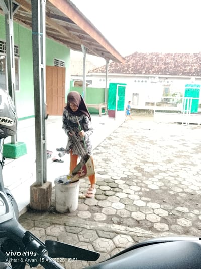 The photo, taken from the perspective of a motorcycle, shows a woman wearing a headscarf sweeping debris into a trash can. The woman is wearing an orange-and-black patterned dress.  The ground is paved with hexagonal stones. There is a small boy in a blue shirt walking away from the camera in the background. The photo was taken in Anjatan, Indonesia, at the coordinates -6.419 latitude and 107.979 longitude.  The image was captured on April 15, 2023 at 15:51.  The woman is in the process of sweeping debris with a broom into the trash can.  The photo was taken in front of a building with a green door and a tiled roof.  There is a motorcycle parked in front of the building.  The building is covered in a green paint.