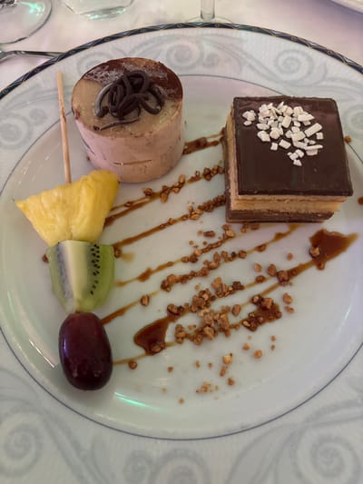 A dessert is presented on a white plate with a blue and white patterned rim. The dessert consists of a cylindrical cake topped with dark chocolate curls, a rectangular chocolate cake layered with cream filling, and a skewer of fruit containing pineapple, kiwi, and a single grape. The plate is decorated with drizzles of caramel sauce and scattered chopped nuts. The location is identified as Vera, Spain.