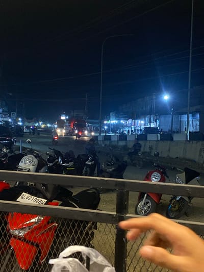 The image depicts a night scene in Tanjongkapai, Indonesia. Motorbikes are parked along a street with buildings illuminated by streetlights and storefronts. Traffic is visible in the distance, indicated by headlights and taillights. A metal fence is present in the foreground, partially obscuring the view. A hand is visible at the right of the image. A license plate BL 62190 BW is visible on one of the motorbikes.