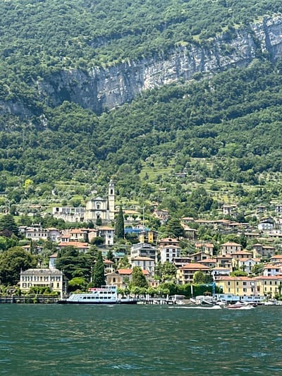 This is a landscape photograph of a coastal town nestled against a steep, verdant mountain. The town, with its characteristic terracotta roofs and colorful buildings, hugs the shoreline. A body of water, possibly a lake or the sea, occupies the right side of the frame, its surface rippling with a deep green hue. The mountain on the left is covered in dense green foliage, with rocky outcrops visible through the trees.
The scene depicts a serene daytime setting, with the bright light suggesting a sunny day. The architecture of the town, with its multi-story buildings, indicates a European location. The presence of a white boat docked at the water's edge suggests a harborside or waterfront community. There are no visible people in the image, focusing the viewer's attention on the natural and built environment. The overall mood is peaceful and picturesque, capturing the charm of a Mediterranean coastal town. No visible text is present in the image.