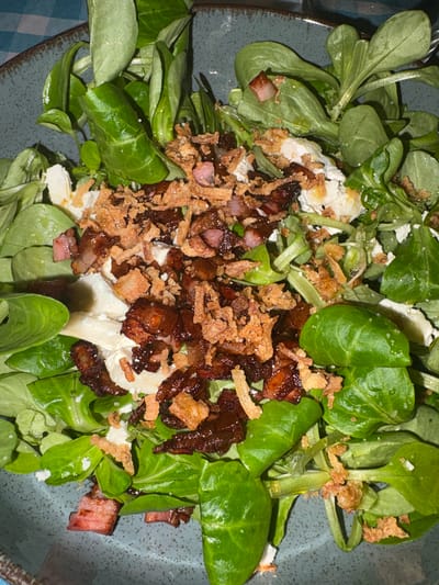 This close-up, overhead shot captures a vibrant salad served in a speckled grey-blue ceramic bowl. The main component is a generous base of fresh, bright green leafy vegetables, consistent with lamb's lettuce or corn salad (Mâche). Scattered across the greens are irregular crumbles of white cheese, possibly feta or goat cheese, and numerous pieces of cooked meat. These meat pieces vary in color from light pink to dark, caramelized brown, strongly resembling diced bacon or ham. Intermingled with the meat are abundant golden-brown, crispy bits, which appear to be fried onions or crunchy croutons, adding texture to the dish. A hint of a blue and white checkered pattern, likely from a tablecloth or napkin, is visible in the lower-left corner of the frame. The scene suggests an indoor dining setting. No people or visible text are present in the image.