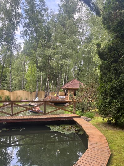 The image depicts a tranquil scene at a private property located in Soloslovo, Russia. A wooden deck and pathway surround a dark body of water, likely a pond or small lake. Lily pads float on the water's surface. In the foreground, a young boy is partially submerged in the water, sitting on an inflatable raft. A wooden fence runs along the perimeter of the property, with trees forming a lush backdrop. A small wooden gazebo with a tiled roof is visible on the far side of the water, connected by a wooden walkway. The overall atmosphere suggests a serene, recreational outdoor setting.