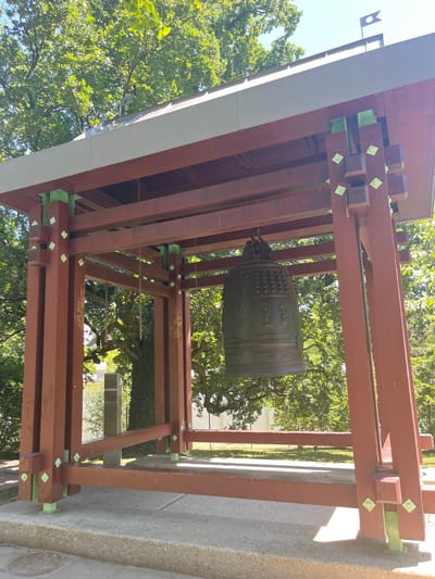 This is an outdoor shot of a large bronze bell suspended from a wooden structure. The bell is ornate, with decorative bands and what appear to be figures or symbols embossed on its surface. The wooden structure is made of thick, dark red beams, some of which are reinforced with bright green metal brackets. The overall construction suggests it is a bell tower or a shrine.

The setting is a park or garden, with lush green trees and foliage visible in the background and above the structure. Sunlight is filtering through the leaves, suggesting it is daytime. The weather appears to be clear and sunny, with dappled light casting shadows. The ground beneath the structure is concrete.

There are no people visible in the image. The activity, if any, is implied by the presence of the bell, which is typically rung for ceremonial or religious purposes. However, the bell appears to be stationary in this image.

Notable details include the contrast between the dark wood and the bright green brackets, as well as the detailed craftsmanship of the bell. There are small green diamond-shaped stickers or markers on some of the wooden beams. The image is taken from a low angle, looking up at the bell and the structure.

The location context of Geneva, Switzerland, combined with the style of the bell and its housing, might suggest a Buddhist temple or a cultural landmark. No visible text is discernible on the bell or the structure.