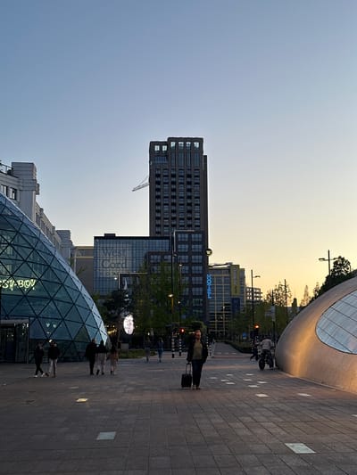 The image captures an urban plaza or street scene in a modern city during the early evening or late afternoon, characterized by a clear sky that transitions from a soft blue on the left to a warm, golden yellow-orange on the right, indicative of sunset or sunrise. Neon signs on buildings are already illuminated, contributing to the dusk ambiance.
In the lower-left foreground, a striking geodesic dome structure made of glass is prominent, reflecting light. Partial text, "S-BOY," is visible on its surface. The ground is paved with large square tiles, alternating in lighter and darker shades.
Towards the right side of the image, a person walks away from the viewer, pulling a black wheeled suitcase. Several other individuals are scattered throughout the mid-ground and background, walking or standing, suggesting a typical bustling city environment.
The architectural backdrop features a mix of contemporary buildings. A tall, dark, rectangular building dominates the mid-left background. Below and slightly to its right, another building displays the text "West Cord." Further to the right, a building features illuminated blue neon signs spelling out "HOTEL" and "Hallo JUMBO," accompanied by a small white logo (possibly an 'M' or 'W'). On the far right, a large, modern building with a curved, metallic facade reflects the warm sky tones. Green trees are interspersed, adding a natural element to the built environment.