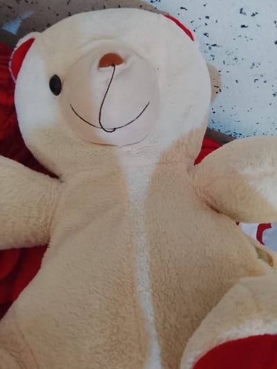 The image shows a close-up, slightly angled shot of a light beige teddy bear. The teddy bear is lying on a red surface, likely a blanket or bed, and appears to be partially leaning against a textured, light blue and grey surface in the background, possibly a wall. The teddy bear has a prominent nose that is a light brown color, and a black eye visible on its left side. Its mouth is a simple black line forming a gentle smile. Red fabric accents are visible on the tops of its ears and possibly on parts of its limbs. The teddy bear's fur is plush and appears to be made of a soft material. The lighting suggests it is indoors, and the overall composition is a still life of the toy. No specific location within Kümüş-Aziz, Kyrgyzstan can be determined from the visual information present in the image.