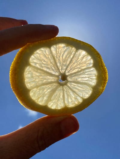 A close-up, low-angle shot captures a translucent slice of lemon held between a person's thumb and forefinger against a bright blue sky. The sunlight shines through the lemon slice, illuminating its segmented interior and yellow rind. The background is a clear sky with a hint of glare from the sun. There are no visible structures or landmarks associated with Красномакское сельское поселение, Ukraine.