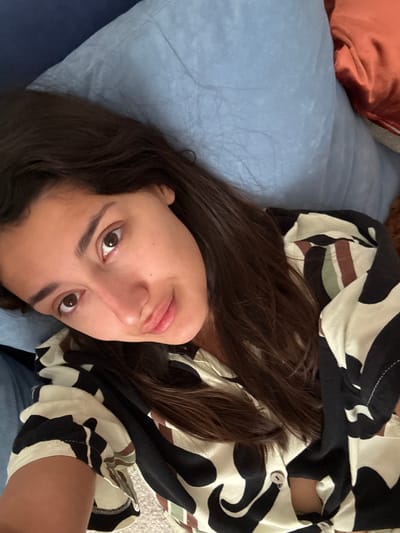 A woman with long brown hair lies on a blue pillow. She wears a patterned shirt with black, white, and brown elements. A rust-colored pillow is visible in the upper left corner of the frame. The image is taken from an overhead perspective. The location is unknown, the request to include Miljevci, Montenegro appears unfounded based on the visual information provided.
