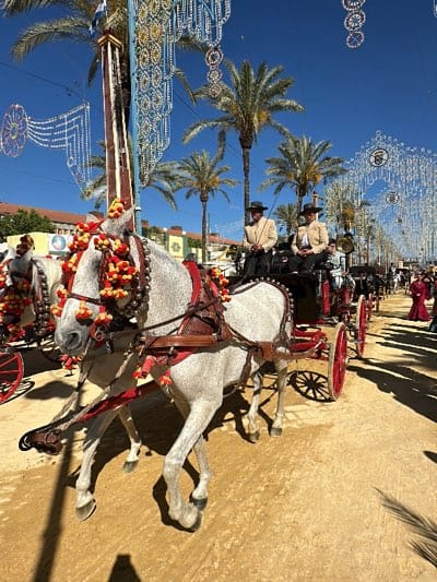 The image shows a horse-drawn carriage procession in Jerez de la Frontera, Spain. Two men dressed in traditional attire are seated in the carriage. The horses are gray and adorned with decorative tassels and bells. Palm trees line the street, and festive lights are strung overhead. Other carriages and people in traditional clothing are visible in the background. The street is a light tan color, likely dirt or sand.