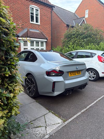This image depicts a static daytime scene on a residential street in Maidstone, United Kingdom, characterized by an overcast sky. The central subject is a light grey BMW M2 coupe (G87 generation), registered with the UK license plate "ST22 VEB" (indicating a 2022 registration). The vehicle is positioned on grey concrete paving slabs, partially obscured on the left by a dense green hedge exhibiting some yellowish leaves. The BMW features black multi-spoke alloy wheels, a black roof, a body-colored rear spoiler lip, and a prominent rear diffuser accommodating four circular exhaust outlets.
Behind the BMW, a white, five-door Ford Fiesta is partially visible, parked on a dark asphalt surface that shows a faded white line along its edge.
To the left of the frame, a red brick house features a white-framed bay window on the ground floor (partially obscured) and a white-framed, arched-top window on the first floor. This house has a brown tiled roof with visible moss accumulation. A black rainwater downpipe is visible on its right side. In the background, a second red brick house is partially visible, displaying a small, high-set rectangular window and a darker, possibly slate-tiled roof. A large green shrub with some reddish foliage stands between the two houses.
No individuals are present. The vehicles are stationary, and the overall scene conveys a typical, quiet residential environment. Weeds are observed growing in the cracks of the concrete paving and along the edge of the asphalt.