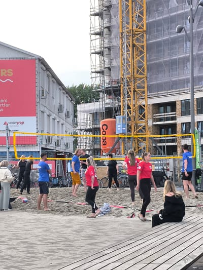 A group of people are playing beach volleyball on a sand court in Alkmaar, Netherlands. A yellow net divides the court. Several individuals are actively engaged in the game, with some positioned on the court and others observing. The background features a building with a large red advertisement, a construction site with a yellow crane, and other urban elements. The ground surface is a paved walkway made of wooden planks. The weather appears to be overcast.