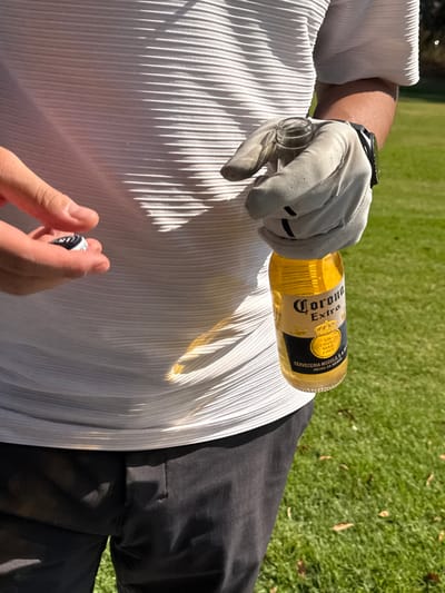A golfer holds a Corona beer bottle resting on their white pleated shirt and black pants. The golfer, wearing a golf glove and a watch, grips the bottle with one hand, while the other holds a golf ball marker. The beer has spilled on the shirt. The background shows green grass, possibly a golf course.