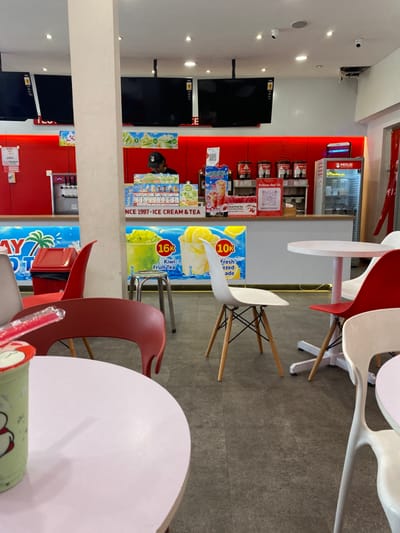 The scene is inside a cafe or restaurant in Kecamatan Piyungan, Indonesia, at the coordinates -7.837 latitude and 110.474 longitude. The cafe has red walls with a white counter separating the customer seating area from the counter. The counter has a sign that reads "INCE 1997 - ICE CREAM & TEA". There are two large, flat-screen televisions mounted on the wall behind the counter. A person, wearing a black cap, stands behind the counter. There are several tables with chairs. One of the tables is positioned closest to the camera, and a half-filled plastic cup containing a green beverage sits on the table. The cup is accompanied by a red plastic straw.
There are four chairs positioned around the table: a red chair with a wooden frame and a white chair with four wooden legs. There are two other chairs: one white and one red, both made of plastic. The chairs are empty. The floor is a mottled grey color.