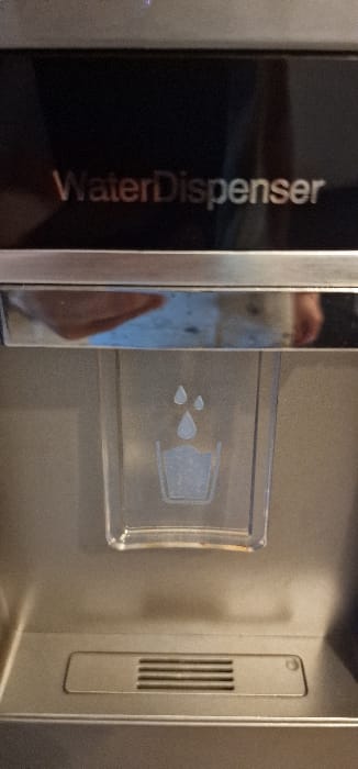 A frontal, close-up image depicts a portion of a modern water dispenser unit in an indoor setting in Blaye, France. The upper section features a dark, reflective panel with the embossed text "WaterDispenser" prominently displayed in a light metallic color. Directly below this, a narrow, highly reflective chrome strip shows blurred, distorted reflections, including what appears to be a human hand or arm and ambient lighting. Centered below the chrome strip is a clear, rectangular plastic component. This component contains an etched or printed graphic: two teardrop shapes representing falling water droplets, positioned directly above a stylized outline of a drinking glass. Below this dispensing icon, near the bottom of the frame, is a rectangular metallic grate with five horizontal slits, likely serving as a drain. The surrounding surface of the dispenser is a brushed metallic finish. No explicit human figures are visible, though reflections suggest proximity to the unit.