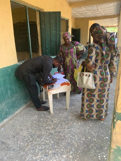 The image shows three people in what appears to be a polling station in Andaha, Nigeria. Two women wearing brightly patterned dresses and head wraps stand near a man who is seated at a small table. The man is dressed in a dark suit and is writing on a piece of paper. Papers and a clipboard are on the table. A handbag rests near the table. The location seems to be outdoors but adjacent to a building with yellow walls and green window frames. The ground is a patterned concrete. Writing is visible on a dark chalkboard-like surface visible through a window.