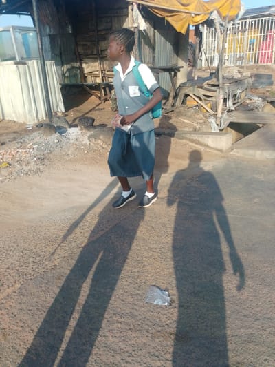 In Garko, Nigeria, a young person in a school uniform, consisting of a grey polo shirt with a crest and blue shorts, walks on a dirt road. The person is carrying a booklet or a small notebook in their right hand. The environment appears to be a market or a roadside stall area, with corrugated metal structures, wooden planks, and a yellow awning visible in the background. The ground is rough and uneven, with scattered debris. The person's shadow stretches long on the road, indicating it is either early morning or late afternoon. Another shadow of an adult figure is also cast on the road. A piece of white plastic trash is on the ground near the shadows.