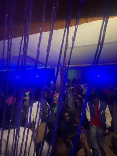 The image captures a dimly lit indoor scene, likely a party or gathering, illuminated by vibrant blue lights. The foreground is partially obscured by dark, rope-like strands, possibly part of a decorative element or curtain. Beyond these, a crowd of people is visible, many in casual attire.
In the mid-ground, several individuals are distinguishable. To the left, a person in a white t-shirt is partially visible. Further to the right, a person wearing a blue and white varsity jacket over a red shirt stands, with grey pants. Another individual in the mid-ground wears sunglasses and a black top. The background shows more indistinct figures and the general ambiance of a lively event, with some people dancing or socializing. The ceiling is visible at the top, featuring a combination of white and wooden elements. The overall impression is one of a social event in a somewhat confined space, characterized by low lighting and a festive atmosphere. The location in Wumat, Nigeria, suggests a cultural context for this gathering.