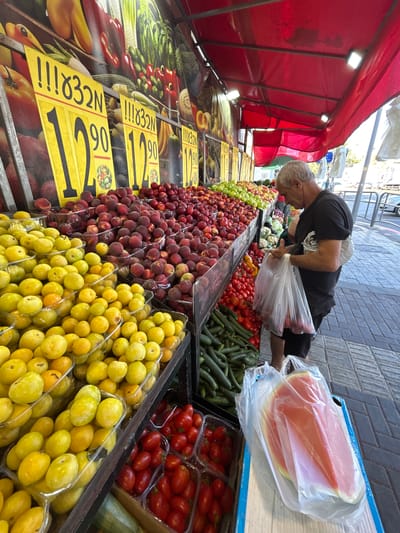 A man with graying hair and a black t-shirt is bent over a fruit and vegetable stand, reaching into a clear plastic bag. He appears to be selecting produce. The stand is outdoors, likely a market, with a red awning overhead. Bright lights are mounted on the underside of the awning, illuminating the goods.
The foreground of the image is filled with a variety of fruits and vegetables displayed in bins and on shelves. Prominent among them are piles of red peaches or nectarines, and a large quantity of yellow, plum-like fruits. Behind these, rows of green cucumbers and red tomatoes are visible. Further back, more vegetables like cauliflower and possibly asparagus can be seen.
Several yellow signs with black text are placed in front of the produce. One sign clearly reads "12.90" with some other unreadable text above it. Another sign has similar pricing information. The overall impression is of a bustling outdoor market scene, likely in Israel given the location context. The time of day appears to be daytime, with bright sunlight suggesting fair weather. The man's focus on selecting produce suggests he is a customer or a vendor preparing to sell. The abundance of fresh produce indicates a vibrant market atmosphere.