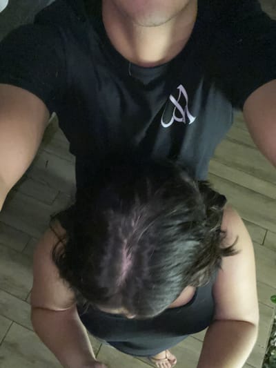 A high-angle photograph captures two individuals in what appears to be an indoor or covered outdoor setting with light-colored, possibly tiled or planked flooring. The person holding the camera is partially visible from the mid-chest upward, wearing a black short-sleeved t-shirt adorned with a white stylized 'M' or similar symbol on the left chest, and a thin chain around their neck. Positioned directly below and in front of the camera-holder is a second individual, viewed from the top of the head to the upper torso. This person has dark, wavy hair with a prominent part revealing some scalp, and wears a dark-colored tank top or dress. Their right foot, wearing a light-colored flip-flop, is visible on the floor. A small patch of green foliage is present on the floor's far right edge. No visual identifiers within the image confirm the location as Coatzacoalcos, Mexico.