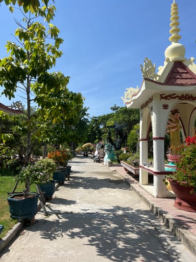 This image shows a pathway in a temple complex in Cam Ranh, Vietnam, on a sunny day. The pathway is paved and lined with potted plants and decorative statues. To the right, a small, ornate pavilion with a pagoda-style roof houses a statue of a multi-armed figure. The pavilion is decorated with red and white accents and intricate carvings. Beyond the pavilion, a garden area is visible, with lush green trees and various flowering plants. Further down the path, there are more statues, including a green dragon and a golden dragon sculpture. Several people are seen in the distance, walking along the path or seated. The sky is clear blue, and the sun casts shadows from the trees and structures onto the ground. The overall impression is one of a serene and well-maintained religious site.