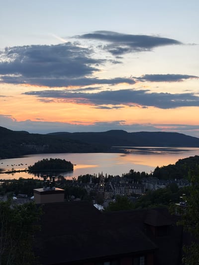 The image depicts a scenic view of Mont-Tremblant, Canada, at dusk. In the foreground, a dark-shingled roof of a building dominates the lower portion of the frame. Above this, a cluster of metallic chimneys stands out against the twilight sky.
The midground reveals a charming village nestled among lush green trees, characterized by its European-style architecture with numerous gabled roofs and dormer windows. A prominent clock tower with a spire marks the center of the village. Beyond the village, a tranquil body of water, likely a lake, reflects the colors of the sunset. A small, tree-covered island sits in the middle of the lake.
The background features rolling hills covered in dark green foliage, receding into the distance. The sky above is a spectacle of color, with hues of orange, pink, and yellow from the setting sun blending into shades of gray and blue from the clouds. The overall impression is one of peacefulness and natural beauty at the end of the day in this picturesque Canadian location.
