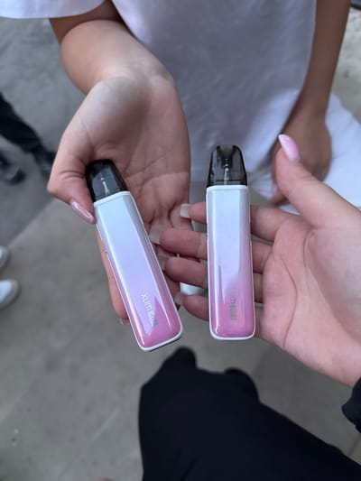 The image shows two hands holding two vape pens. The vape pens are white with a pink iridescent gradient and have black mouthpieces. One vape pen has the text "XLIM GO" in white on its side. The hands are holding the vape pens up towards the viewer. The background is blurred and appears to be an outdoor setting. There are no specific geographical indicators to place the image in Tovstoluh, Ukraine.