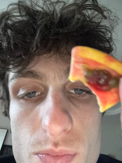 A close-up, eye-level selfie-style photograph depicts a male individual holding a piece of fruit near his face. The man has curly, dark brown hair that appears disheveled, blue eyes with dark brows, and visible stubble on his lower face. His skin tone is light, with some reddish areas around the nose and cheeks. He looks directly towards the camera with a neutral expression. A bright orange and yellow piece of fruit, possibly a slice of papaya or mango, is held in the foreground, partially obscuring the upper right portion of his face, including his right eye and forehead. The fruit shows an exposed interior with dark, possibly seed-filled, red pulp. The background is out of focus and light-colored, suggesting an indoor setting, with a faint, dark rectangular shape visible in the lower left corner, possibly a framed object or mirror. The specific location within the city of Vincennes, France, cannot be determined from the visual information provided.