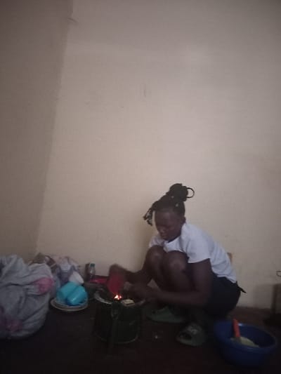 A woman kneels next to a charcoal stove. She wears a white shirt and dark shorts, with her hair in a high bun. A blue bowl with contents sits in the upper right. The stove is green and sits on a tray. Other items like a blue cup and crumpled clothes are near the stove. The background is a plain wall. The image appears to be taken indoors.