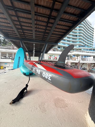 The image shows two paddleboards resting on a sandy surface, likely a beach. The paddleboards are oriented vertically in the frame. The larger paddleboard is red and gray with black accents, and the text "MYBOAT 380 84 length" is visible on its side. It has a black fin attached to its underside. A coiled black leash extends from the fin to a black strap on the ground. A smaller, light blue paddleboard is partially visible beneath the larger one, with the text "MYBOAT" also visible on its side.
In the background, a modern, wavy-facade building with many balconies rises high into the sky. The sky appears overcast, suggesting a cloudy day. The setting appears to be a resort area, with what looks like lounge chairs and umbrellas visible behind the paddleboards and near the building. The overall impression is a relaxed, possibly vacation-oriented scene. No people are clearly visible in the image. The lighting suggests it is daytime. The location cues point to a seaside or waterfront area, with the building and lounge chairs typical of a resort.