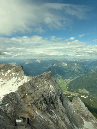 This image is a wide, high-angle, vertical shot of a rugged mountain landscape. In the foreground, a steep, rocky mountain face dominates the left side of the frame, with patches of snow clinging to its crevices. A small, dark, hut-like structure is visible nestled into the rock face near the bottom left. The mountain slopes downwards into a verdant valley dotted with buildings and trees, indicative of a town or village. This valley is framed by layers of receding mountain ranges that stretch into the distance. The sky above is a vibrant blue, filled with scattered, fluffy white clouds. The scene depicts a dramatic natural environment with signs of human habitation in the valley below. The viewpoint suggests the photo was taken from a very high elevation, possibly a mountaintop or a cable car, looking down and across the valley. The presence of a town in the valley and the distinct mountain ranges are consistent with the location of Ehrwald, Austria.