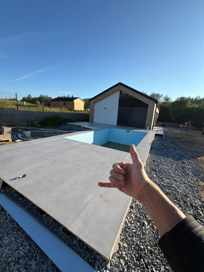 An exterior, wide-angle, eye-level shot shows a partially constructed pool area in Horní Planá, Czechia. A person's arm and hand are in the foreground, giving a "shaka" gesture, with their thumb up. The pool is empty, with light blue walls, and a dark green substance at the bottom. The pool is surrounded by a tiled patio. In the background, a modern house with a wooden facade is visible, along with a gravel-covered yard and some greenery. A clear blue sky with a thin white cloud stretches overhead.
