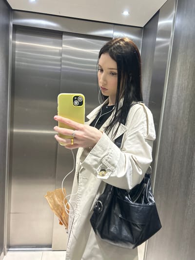 A young female is depicted taking a mirror selfie inside an elevator in Cachan, France. She has long, dark hair and is wearing a light beige trench coat over a dark top. White wired earphones are visible in her ears. Her left hand holds a bright yellow smartphone, which features a dual-camera system. On her left hand, a gold-colored band is visible on her index finger and a silver-colored ring with a clear stone is on her middle finger. A black, semi-glossy shoulder bag with silver-colored rings and a zipper is worn over her right shoulder. A crumpled brown paper bag is partially visible, held in her left hand below the smartphone. The background features brushed metal elevator doors and a dark, possibly textured, side wall. Overhead lighting reflects in the metallic surfaces.