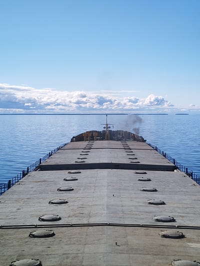 The image depicts a view from the deck of a large freighter ship at sea. The deck is gray and industrial, with numerous circular hatches arranged in parallel lines. A low railing runs along the edge of the deck. In the distance, the calm blue water meets a horizon lined with clouds and a few small islands. The sky is mostly blue with scattered clouds. A plume of black smoke rises from the ship's superstructure, which is visible further down the deck. The location cannot be determined from the image.