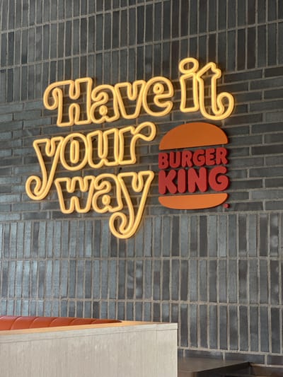 The image shows a Burger King logo and neon sign on a dark grey brick wall. The neon sign spells out "Have it your way" in a yellow glow. The Burger King logo is a red sign shaped like a hamburger bun with "BURGER KING" written in red capital letters inside. The wall is made of vertically laid rectangular bricks. In the foreground, the top of a light-colored wooden counter with an orange upholstered seat is visible. The location is identified as Dub, Montenegro. There are no people or animals visible in the image.
