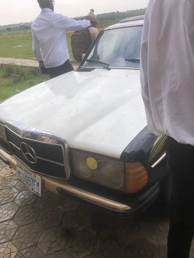 This media file depicts two individuals and a white Mercedes-Benz car, viewed from a slightly elevated, tilted perspective. The scene appears to be outdoors, possibly in a paved area with a grassy field and distant trees. The weather seems to be rainy, as indicated by water droplets on the car's hood and a wet appearance on the ground.
One person, wearing a white shirt, is visible near the car's open driver-side door, facing away from the camera and towards the grassy field. Another person, partially obscured by a white garment, is standing near the car's hood.
The car is a classic Mercedes-Benz model, identifiable by its distinct grille and headlights. A license plate is visible on the front of the car, with the text "MGH-657FM" and "PLATEAU". The overall impression is that of a casual outdoor setting, potentially in Nigeria given the "PLATEAU" text on the license plate, which could refer to Plateau State. The overcast sky and wet conditions suggest it might be a cloudy or rainy day.