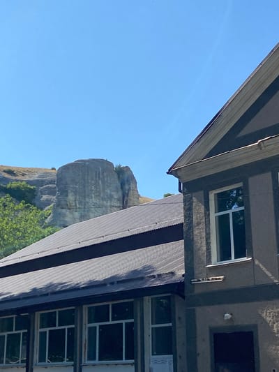The image shows a view of a rocky outcrop and some buildings under a clear blue sky. In the foreground, to the right, is a part of a building with a dark gray facade, a pitched roof with dark gray shingles, and a white-framed window. To the left of this building, another building with a similar dark gray shingled roof is visible, featuring multiple windows with white frames. In the background, a large, weathered rock formation with vertical striations stands on a grassy hillside. Trees and bushes are present on the lower slopes of the hill. The overall scene appears to be outdoors in a rural or semi-rural setting.