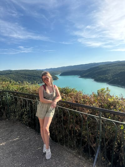 A woman stands on a gravel pathway, leaning against a dark metal railing. She is wearing a floral print romper, sunglasses, and white sneakers. Behind her, a large turquoise lake is surrounded by lush green hills. The sky above is a clear blue with scattered white clouds. The location is the Lac de Vouglans, a reservoir located in the Jura department of France, near the commune of Moirans-en-Montagne.