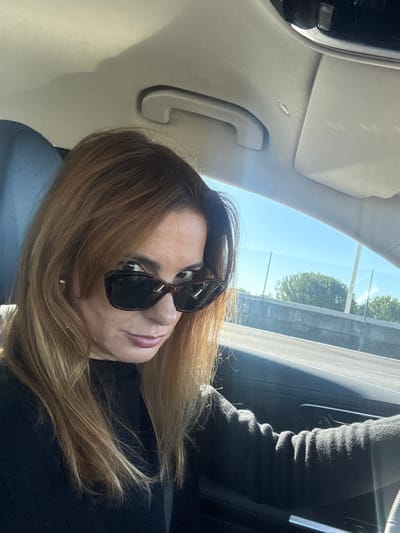 A single adult female is depicted seated in the driver's position of a vehicle, in the city of Alcochete, Portugal. She possesses long, auburn-to-light-brown hair with lighter highlights, styled with a frontal fringe. She is wearing dark, rectangular-framed sunglasses and a dark, long-sleeved garment, appearing to be a sweater. A small, circular gold-colored earring is visible on her right earlobe. Her head is slightly tilted, and she is looking directly at the camera with a neutral facial expression. The interior of the vehicle shows a light-colored headliner and a grey overhead grab handle on the passenger side. Outside the vehicle, visible through the window, is a clear blue sky above a concrete road barrier. Behind this barrier, a metal mesh fence and green trees are present. The scene is illuminated by strong sunlight entering from the right side of the frame.