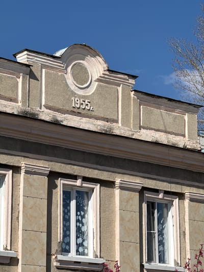 The image shows the corner of a building in Borzya, Russia. The architectural style features ornate detailing and a circular design with the number "1955" inscribed on the facade. The building's exterior is predominantly gray with lighter accents, and its condition shows signs of age and weathering. Adjacent to the corner are windows decorated with balloon and star designs. The sky is blue with sparse tree branches visible at the top of the frame. The photo is oriented on an angle, with the building appearing to lean diagonally.