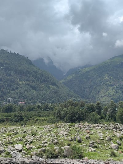 The image captures a panoramic view of a mountainous landscape with lush green vegetation and a cloudy sky. In the foreground, a rocky terrain is dotted with scattered stones and patches of grass. Beyond this, a dense forest covers the slopes of the mountains, with some buildings visible nestled among the trees at the lower elevations. The middle ground features more densely forested hills, gradually receding into the distance. The background is dominated by imposing mountains, partially obscured by heavy, overcast clouds, suggesting a possibly cool or misty atmosphere. One notable building, a large red-roofed structure, stands out amidst the greenery on a mountainside. The scene is characteristic of the mountainous regions of India, specifically the area around Bahang, known for its natural beauty and proximity to the Himalayas.