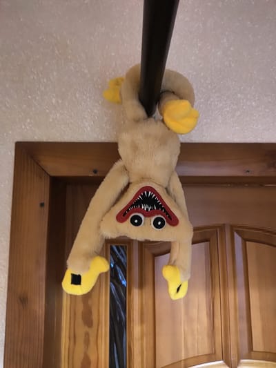 The image shows a fluffy, light-tan plush toy resembling a monster with a large, sharp-toothed mouth and two black eyes outlined in red. The toy has yellow, mitten-like hands and feet, with black squares on the palms and soles. It is draped over a dark, cylindrical object, possibly a pole or rod. The background features a textured, light-colored wall on the left and a wooden door frame on the right. The door is made of several panels and has a distinct wood grain. The lighting suggests an indoor setting, possibly during the daytime. The plush toy appears to be positioned mid-air, suggesting it's either hung or held in place. There is no visible text in the image.