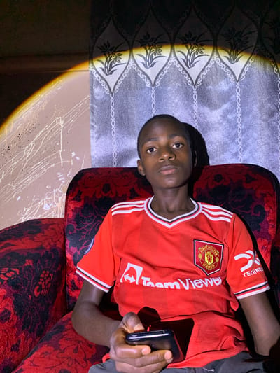 The image captures a young person seated on a red, patterned armchair in Barakin Sabawa, Nigeria. The individual is wearing a red Manchester United jersey with the TeamViewer logo and a Manchester United crest on the chest, and a "TechNow" logo on the sleeve. They are holding a black smartphone in their right hand. The background features a wall with a patterned wallpaper, and a distinct arc of light, likely from a window or lamp, illuminates the scene from the left, casting shadows. The wall to the left of the armchair has a textured finish with white and black splatters. The lighting creates a dramatic effect, with some areas of the image in shadow and others brightly lit.