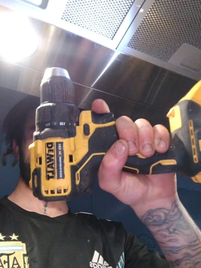 A close-up, first-person perspective shot shows a hand holding a yellow and black DeWalt drill. The drill has a silver chuck and black grip. The hand is partially visible, with dirty fingernails. Behind the drill, a person's face is partially obscured by the drill. They have dark hair and a beard. The person is wearing a black t-shirt with white stripes on the sleeves and a crest on the left chest. A tattoo is visible on the person's neck, and another tattoo, appearing to be a cloudy design, is on their forearm. The background appears to be indoors, with a stainless steel range hood and light fixtures visible. The scene is lit by a bright overhead light source. There is no indication of the location being in Crauthem, Luxembourg.