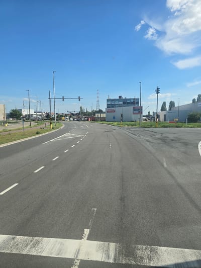The image shows a view from a vehicle looking towards a road intersection under a clear blue sky with some scattered clouds. The road is asphalt with white lane markings and a pedestrian crossing. On the left, there is a green grassy area with a bicycle path and streetlights. In the background, several industrial buildings are visible, including a building with the "EWSGROUP" logo and another with "HANSALFLEX" signage. There are also traffic lights, utility poles, and wind turbines in the distance. The scene appears to be in an industrial area. Based on the visible signage such as "EWSGROUP", this location is likely within the port of Antwerp, Belgium, specifically in an area like the Waaslandhaven.