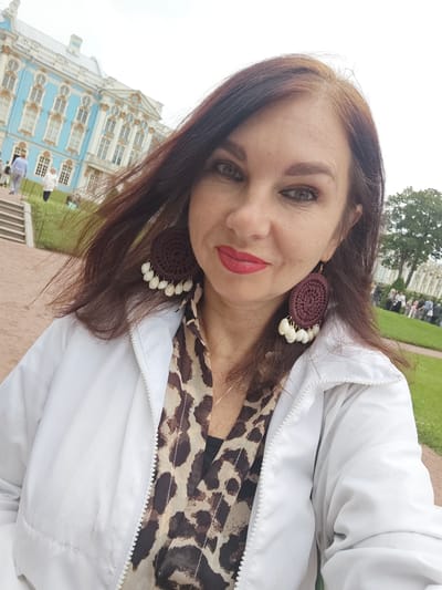 This is a selfie of a woman with medium-length brown hair and distinctive large, round, maroon crochet earrings with white bead accents. She is wearing a white zippered jacket over a leopard-print top and a silver necklace. Her makeup is noticeable, with red lipstick and eye makeup. Her expression appears to be a slight smile.
In the background, a large, ornate blue and white building with numerous windows and decorative elements is visible. This is the Catherine Palace in Pushkin, Russia. Several people are scattered around the grounds, some walking on a path and steps, others gathered on a grassy area. The overall setting suggests a daytime visit to a historical landmark. The weather appears to be overcast.