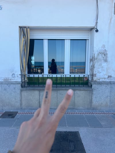 The image shows a building exterior in Sant Pol de Mar, Spain. The wall is partially white with weathered patches, and partially gray. A window with a white frame and closed white roll-down blinds is visible. A striped awning is partially rolled up below the window. A black metal railing sits outside the window. A person's hand is in the foreground, displaying a peace sign. The reflection in the window shows a silhouetted figure inside the building. A gray tiled sidewalk with a small, dark metal utility access cover (Telefonica) is beside the building. The overall setting is outdoors in daylight.