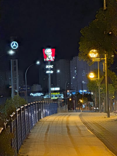 The image depicts a street scene at night, likely in Finestrat, Spain. The perspective is from a slightly elevated position, looking down a curved pedestrian walkway lined with textured concrete and metal railings. The walkway is illuminated by streetlights and the glow from various illuminated signs.

Prominently visible in the mid-ground is a brightly lit KFC sign featuring the iconic Colonel Sanders logo, accompanied by text reading "KFC" and "AUTO". Other signs are also visible, including "Conforama", "vegan", and several others with text that is partially obscured or difficult to read due to the angle and lighting. There is also a stylized illuminated logo that appears to be for Mercedes-Benz.

In the background, several multi-story buildings are visible, with many windows lit, suggesting an urban or semi-urban residential area. Trees with yellow and green foliage are present on the left side of the image, casting shadows and adding to the nighttime ambiance. The overall mood is one of a commercial area active into the evening, with artificial lights dominating the scene. The time of day is clearly night. The weather appears to be clear, as there are no visible signs of rain or fog.