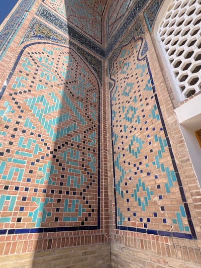 The image shows a close-up of the exterior of a building in Samarkand, Uzbekistan, featuring intricate tilework and brickwork. The wall is adorned with a geometric pattern of small, rectangular tiles in various shades of blue, black, and beige, set against a backdrop of reddish-brown brick. Some sections of the wall are highlighted by the sun, creating a contrast with the shadowed areas. In the upper right portion of the image, a section of a white, honeycomb-patterned lattice is visible, likely part of a window or decorative screen. The overall impression is one of ancient architectural artistry, with the textures of the tiles and bricks clearly discernible.