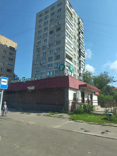 The image captures a street scene in Zhukovsky, Russia, dominated by a tall, grey, multi-story apartment building and a smaller brick building with a red roof in the foreground. A sign above the smaller building reads "РАМЕНСКИЙ ДЕЛИКАТЕС" in green lettering. Two circular signs with a green 'P' are mounted on the apartment building. A blue bus stop sign is visible to the left of the frame. A woman with blonde hair, wearing a black t-shirt and grey pants, walks on the sidewalk carrying a pink shopping bag. Another person, a man wearing a grey t-shirt, stands on the porch of the smaller building. The ground is paved with asphalt, and there is a grassy area with bushes and trees. The sky is blue with scattered clouds. The scene appears to be a bright, sunny day.